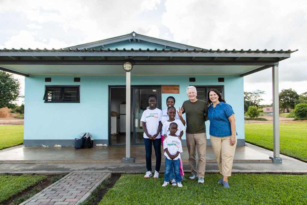 A white couple with three African children smiling in front of a newly built home.